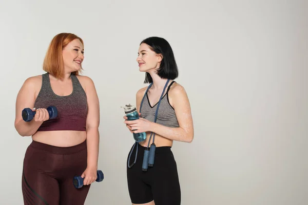 Smiling body positive sportswomen holding jump ropes and sports bottles isolated on grey — Stock Photo