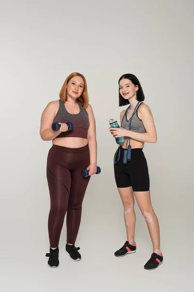 Junge körperlich positive Sportlerinnen mit Sportflasche und Hanteln blicken in die Kamera auf grauem Hintergrund — Stockfoto