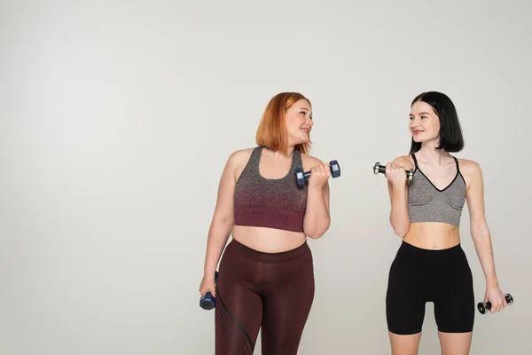 Cheerful body positive sportswomen training with dumbbells isolated on grey — Stock Photo