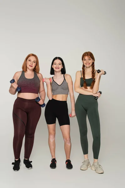 Deportistas sonrientes con cuerpo positivo sosteniendo mancuernas sobre fondo gris - foto de stock