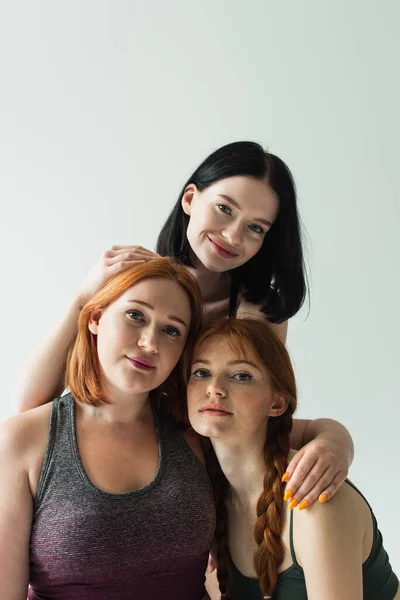 Smiling sportswoman with vitiligo hugging body positive friends and looking at camera isolated on grey — Stock Photo