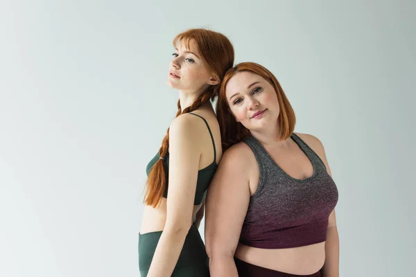 Plus size and freckled sportswomen standing back to back isolated on grey — Stock Photo