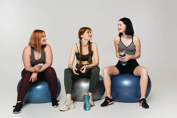 Body positive sportswoman with sports bottle looking at friends on fitness balls on grey background — Stock Photo