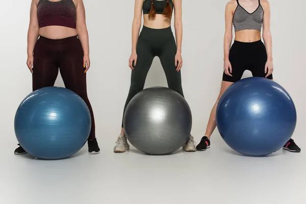 Cropped view of body positive sportswomen standing near fitness balls on grey background — Stock Photo