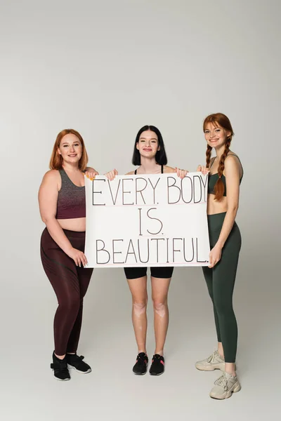 Sportswoman with vitiligo holding placard with every body is beautiful lettering near friends on grey background — Stock Photo