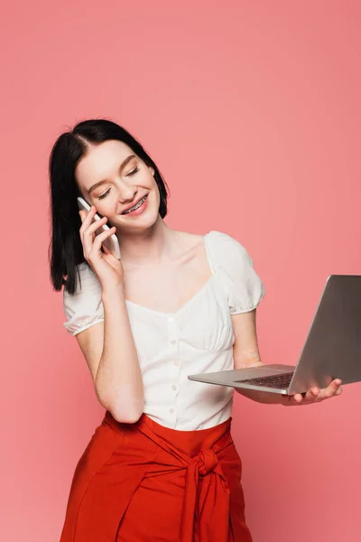 Mujer sonriente con vitiligo hablando por teléfono móvil y sosteniendo portátil aislado en rosa - foto de stock