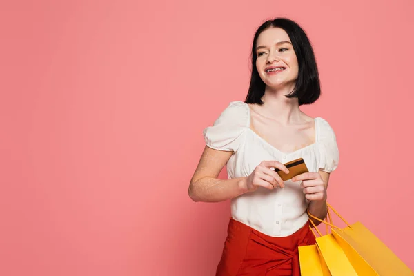 Mujer feliz con vitiligo con tarjeta de crédito y bolsas aisladas en rosa - foto de stock