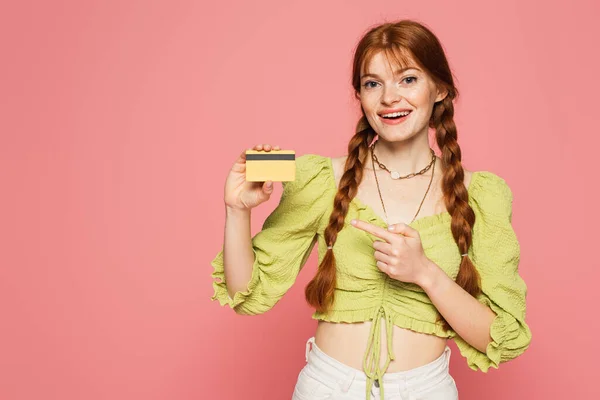 Smiling redhead woman pointing at credit card isolated on pink — Stock Photo
