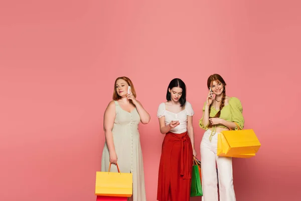 Стильное тело позитивных женщин с сумками, говорящих на смартфонах на розовом фоне — стоковое фото