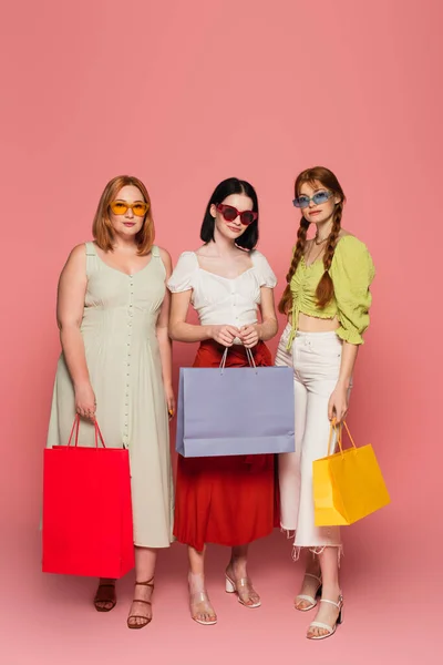 Cuerpo amigos positivos en gafas de sol sosteniendo bolsas de compras sobre fondo rosa - foto de stock