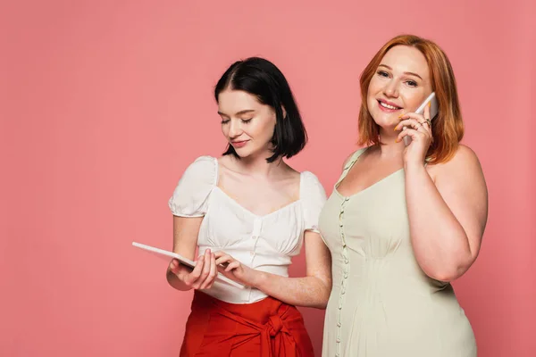 Sonriente mujer de tamaño grande hablando en smartphone cerca de amigo con vitiligo utilizando tableta digital aislada en rosa - foto de stock