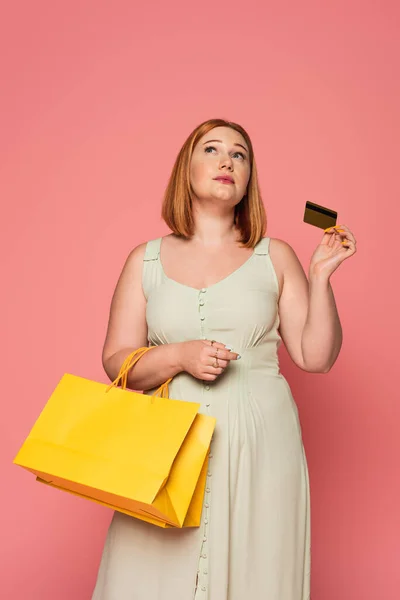 Mujer pensativa más tamaño con tarjeta de crédito y bolsas de compras aisladas en rosa - foto de stock