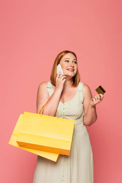 Lächelnde Plus-Size-Frau spricht auf Smartphone und hält Kreditkarte und Einkaufstasche isoliert auf rosa — Stockfoto