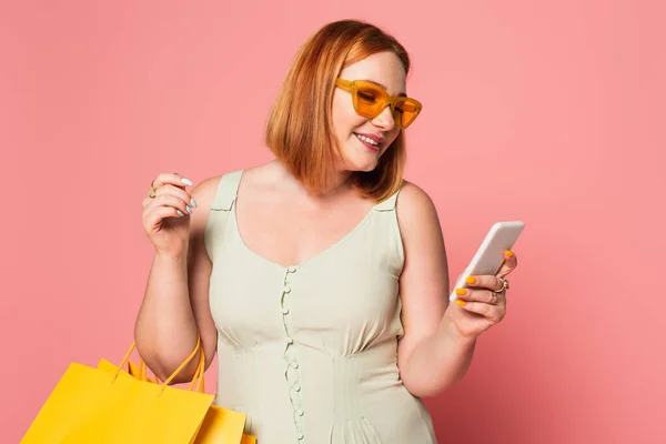 Smiling plus size woman in sunglasses using smartphone and holding shopping bags on pink background — Stock Photo