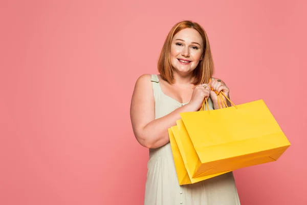 Sonriente mujer pelirroja con bolsas de compras mirando a la cámara aislada en rosa - foto de stock