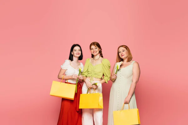 Amigos sonrientes con bolsas de compras sosteniendo flores de calla aisladas en rosa - foto de stock