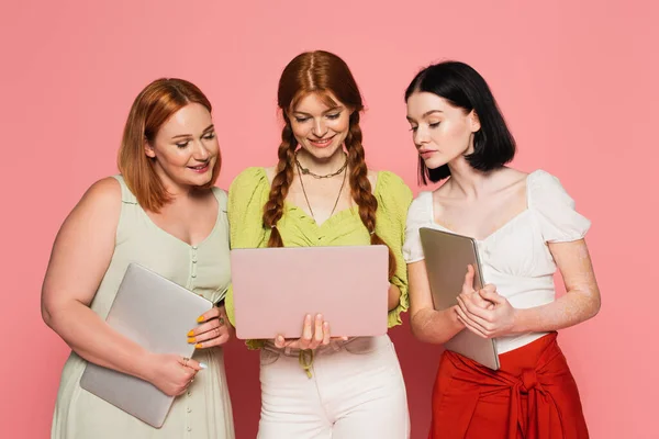 Mulheres sorridentes olhando para amigo usando laptop no fundo rosa — Fotografia de Stock