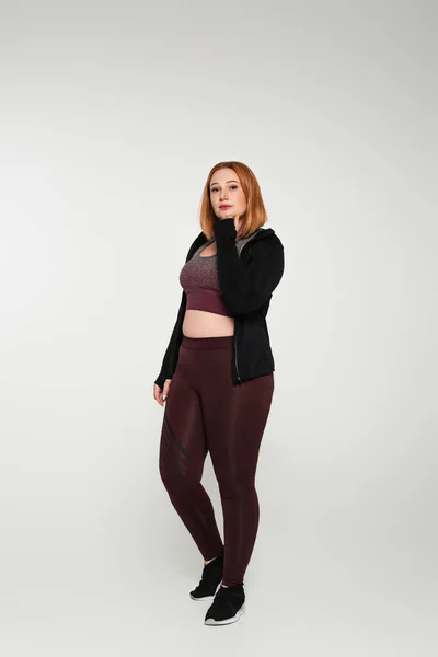 Rothaarige Plus-Size-Frau in Sportbekleidung steht auf grauem Hintergrund — Stockfoto