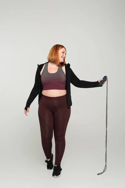 Side view of smiling body positive sportswoman holding jump rope on grey background — Stock Photo