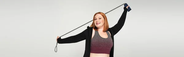 Red haired sportswoman holding jump rope isolated on grey, banner — Stock Photo