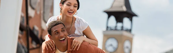 Pretty asian woman smiling while piggybacking on african american boyfriend, banner — Stock Photo