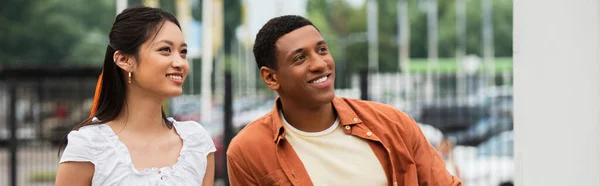 Joven pareja multiétnica sonriendo mientras mira al aire libre, pancarta - foto de stock