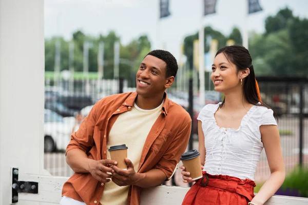 Elegante y feliz pareja multiétnica sosteniendo café para ir mientras mira al aire libre - foto de stock