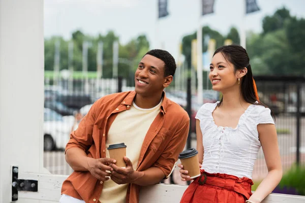 Élégant et heureux couple multiethnique tenant café pour aller tout en regardant loin à l'extérieur — Photo de stock