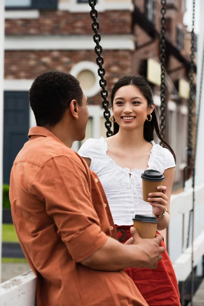 Joyful asian woman with takeaway drink smiling at african american man on city bridge — Stock Photo