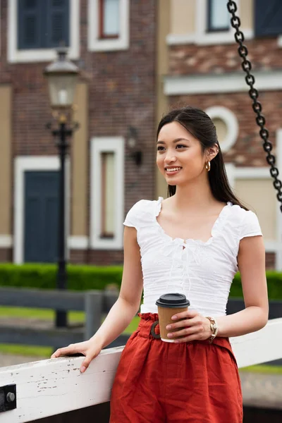 Pretty asian woman with paper cup smiling on bridge in city — Stock Photo