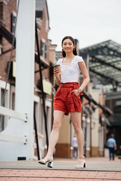 Alegre asiático mulher segurando café para ir enquanto andando no urbano rua com mão no bolso — Fotografia de Stock