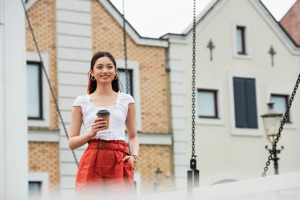 Souriant asiatique femme avec café pour aller debout avec la main dans la poche de shorts à l'extérieur — Photo de stock