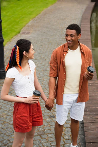 Alegre interracial pareja con takeaway beber cogido de la mano y mirando uno al otro - foto de stock