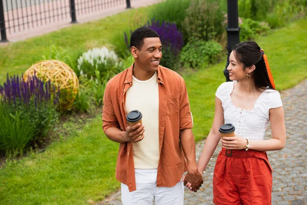 Couple multiethnique joyeux avec café pour aller se tenir la main tout en marchant dans le parc — Photo de stock