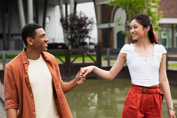 Alegre pareja multiétnica mirándose y tomándose de la mano mientras caminan cerca del agua - foto de stock