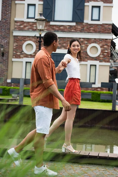 Веселая азиатская женщина, держащаяся за руки с африканским американским парнем во время прогулки по городу — стоковое фото