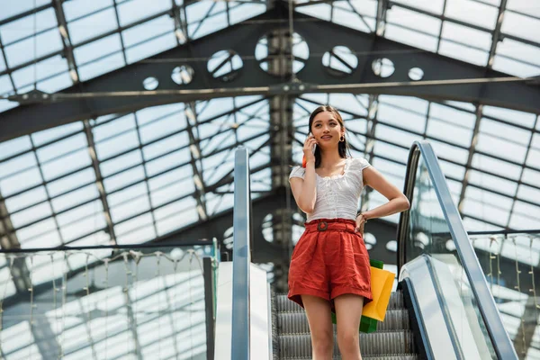 Bonita mujer asiática con bolsas de compras hablando en smartphone en escalera mecánica - foto de stock