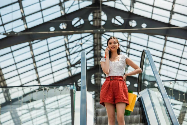 Mulher asiática bonita com sacos de compras falando no smartphone na escada rolante — Fotografia de Stock