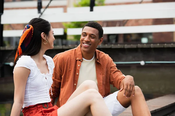 Cheerful interracial couple talking while sitting on street border in city — Stock Photo