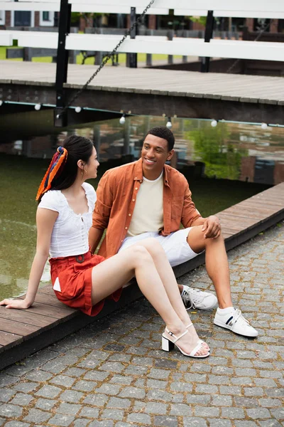 Jovem afro-americano homem sorrindo para asiático mulher na fronteira perto de água — Fotografia de Stock