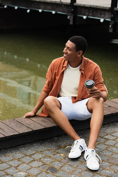 Щасливий афроамериканський чоловік з виносним напоєм, що сидить на кордоні біля води на міській вулиці — стокове фото