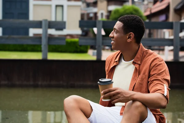 Young african american man holding paper cup while sitting near water on city street — Stock Photo