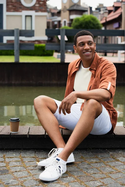 Hombre afroamericano feliz sentado en la frontera cerca del agua y el café para ir en la calle urbana - foto de stock