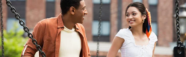 Jovem e feliz casal interracial sorrindo uns para os outros ao ar livre, banner — Fotografia de Stock