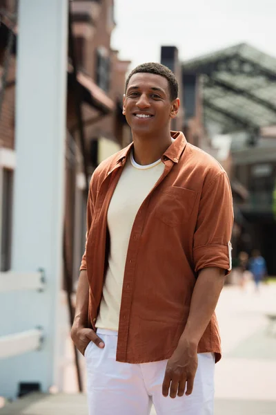 Hombre afroamericano feliz de pie con la mano en el bolsillo en la calle de la ciudad - foto de stock