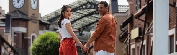 Feliz casal interracial olhando para a câmera enquanto segurando as mãos ao ar livre, banner — Fotografia de Stock