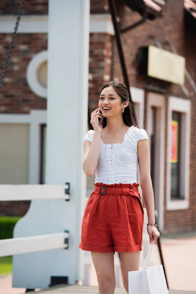 Fröhliche Asiatin telefoniert mit Smartphone, während sie mit Einkaufstasche auf Brücke geht — Stockfoto