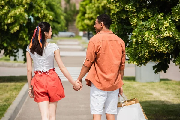 Vista trasera de pareja interracial en pantalones cortos tomados de la mano mientras camina por el callejón - foto de stock