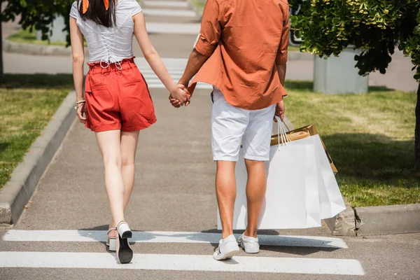 Rückansicht eines beschnittenen multiethnischen Paares in Sommerkleidung, das sich beim Stadtspaziergang an den Händen hält — Stockfoto