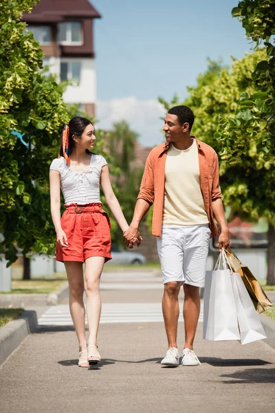 African american man with shopping bags holding hands with asian woman on green alley — Stock Photo